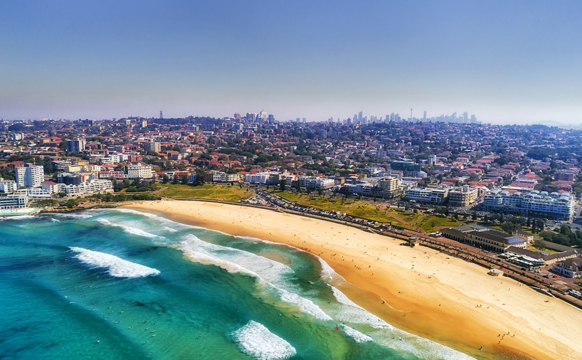 Wave breaks and surf on clear sand of famous Australian Sydney Bondi beach in aerial view with city CBD in background.
