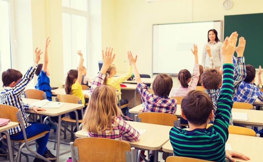 education-elementary-school-learning-and-people-concept-group-of-school-kids-with-teacher-sitting-in-classroom-and-raising-hands