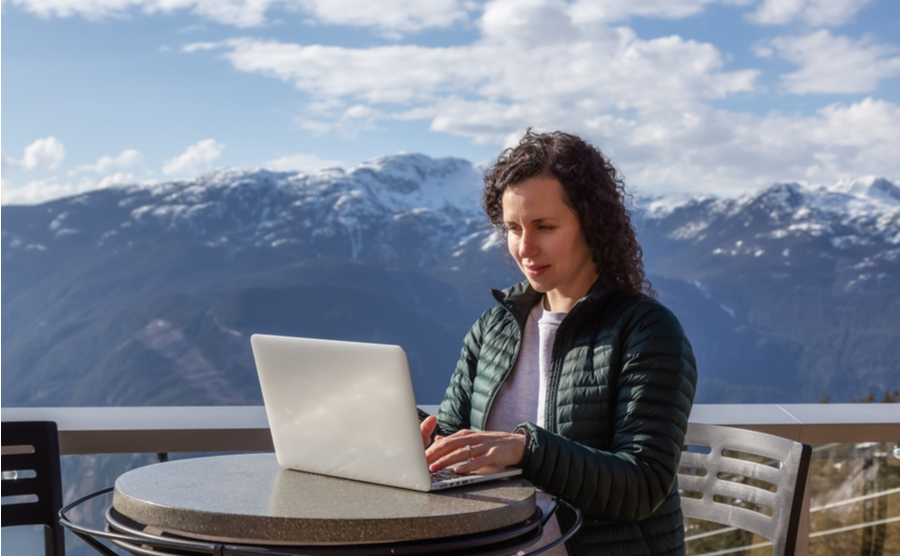 Squamish, BC, Canada - March 9, 2023: Caucasian Woman working on Laptop outside with mountain landscape in background. Sea to Sky Gondola. Adventure Travel